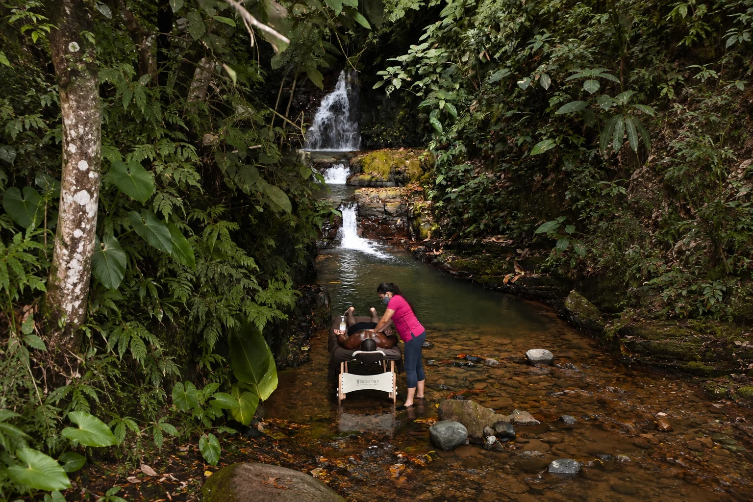 Guest enjoying a riverside massage beneath a tropical waterfall at a boutique jungle hotel in Costa Rica, surrounded by lush rainforest, cascading water, and natural rock pools.
