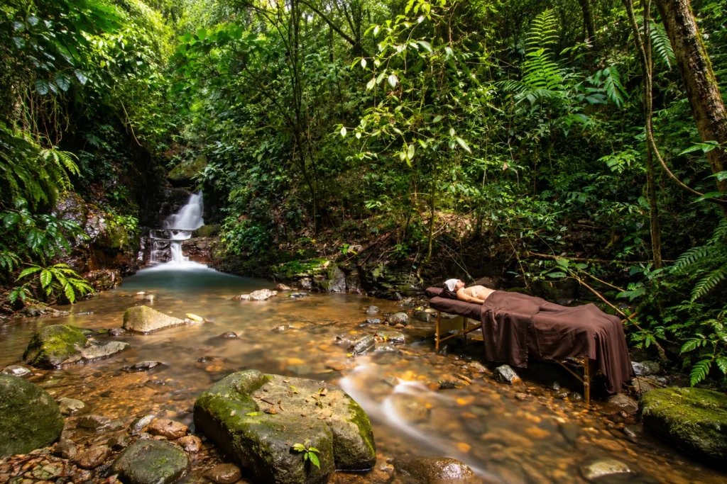 A person lying draped in a brown towel on a massage table positioned directly alongside a clear jungle stream, with a cascading waterfall and lush rainforest foliage in the background.